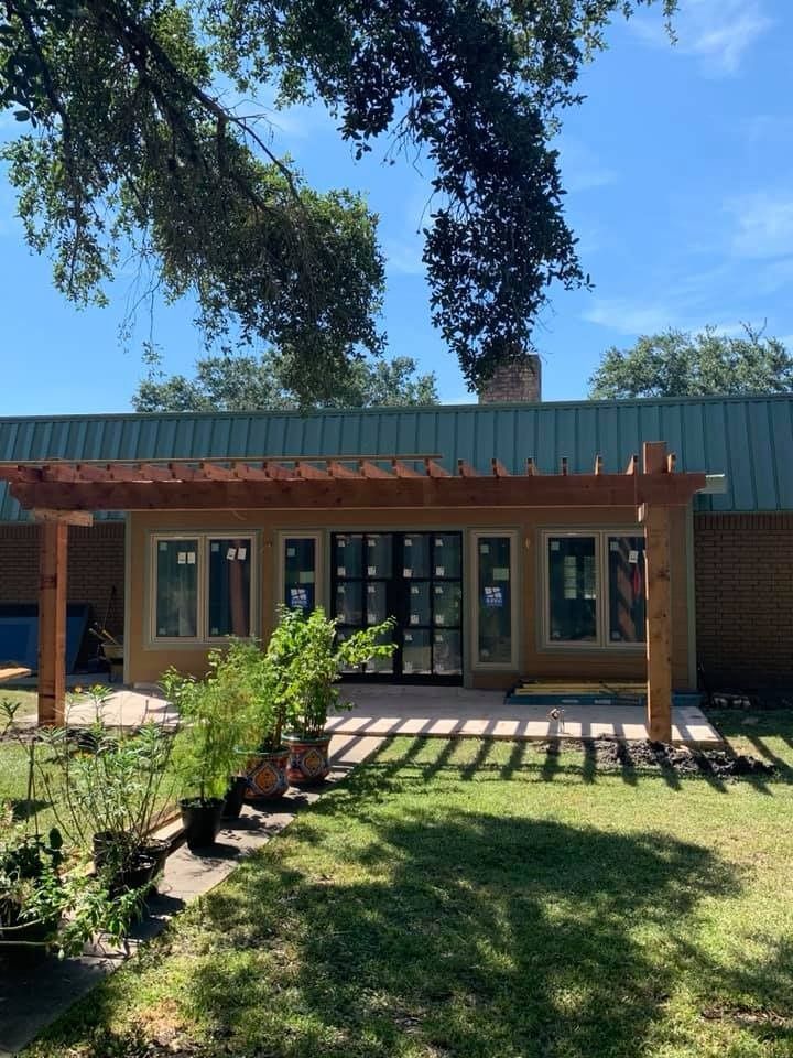 A new wooden pergola stands over a patio in front of a tan house with a green metal roof under a bright blue sky.