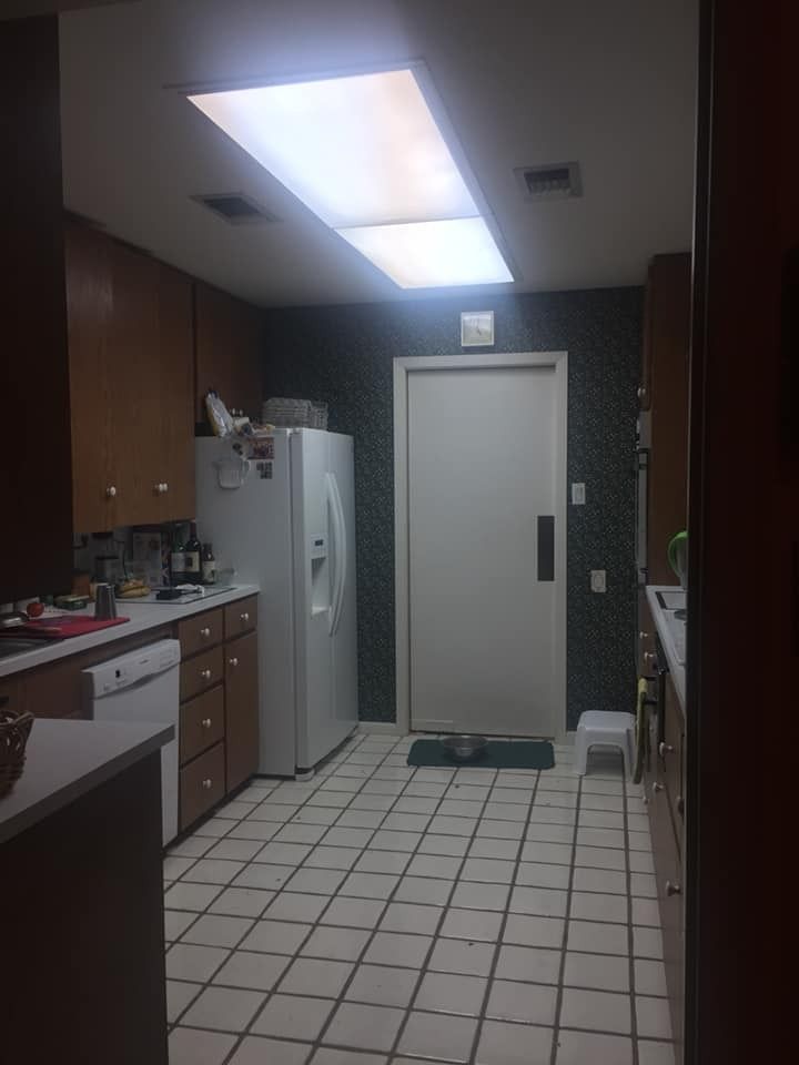 A kitchen with brown cabinets, white appliances, tiled flooring, and a large ceiling light fixture above a doorway.