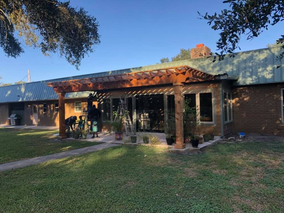 People work on a patio under a large wooden pergola in front of a house with a green metal roof.