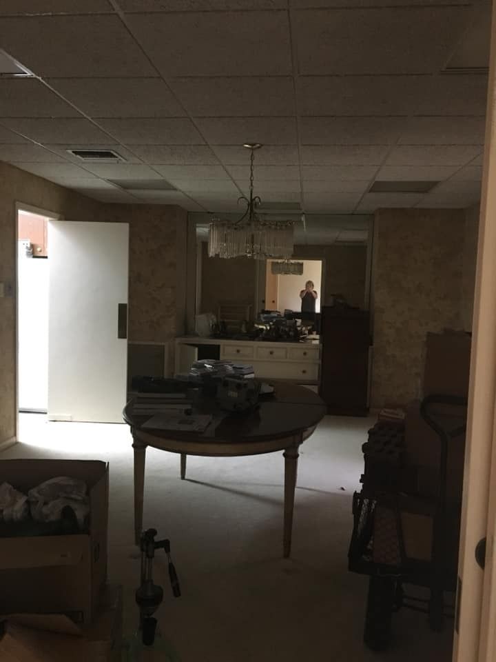 A dimly lit room with a round wooden table, white dresser, and a door leading to a bright exterior area.