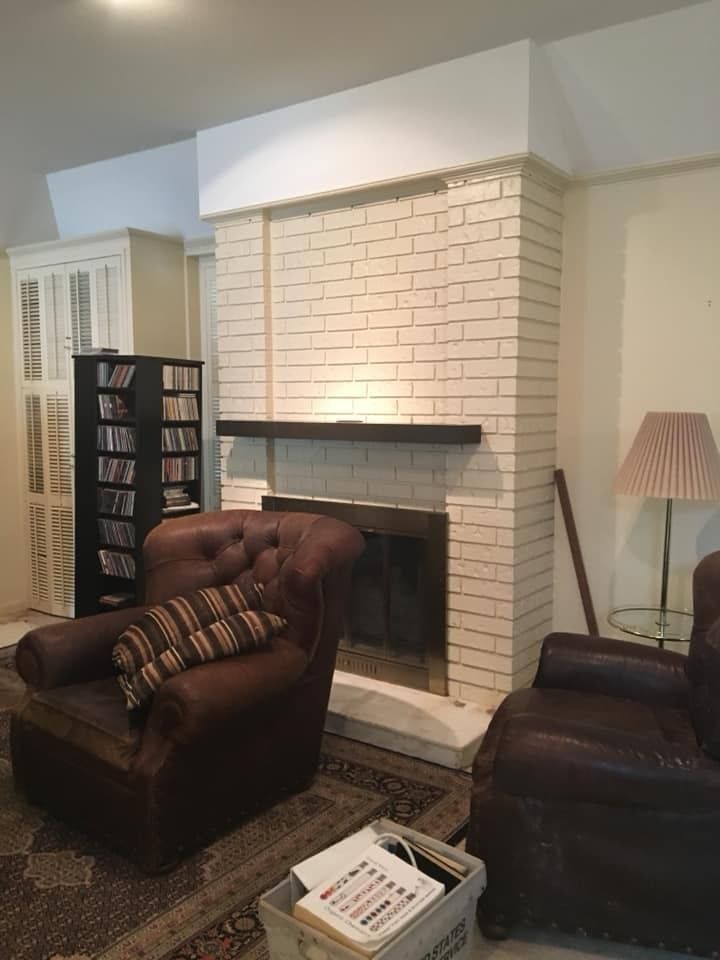 A room with a white-painted brick fireplace featuring a dark mantel, two leather armchairs, and a black CD storage shelf.