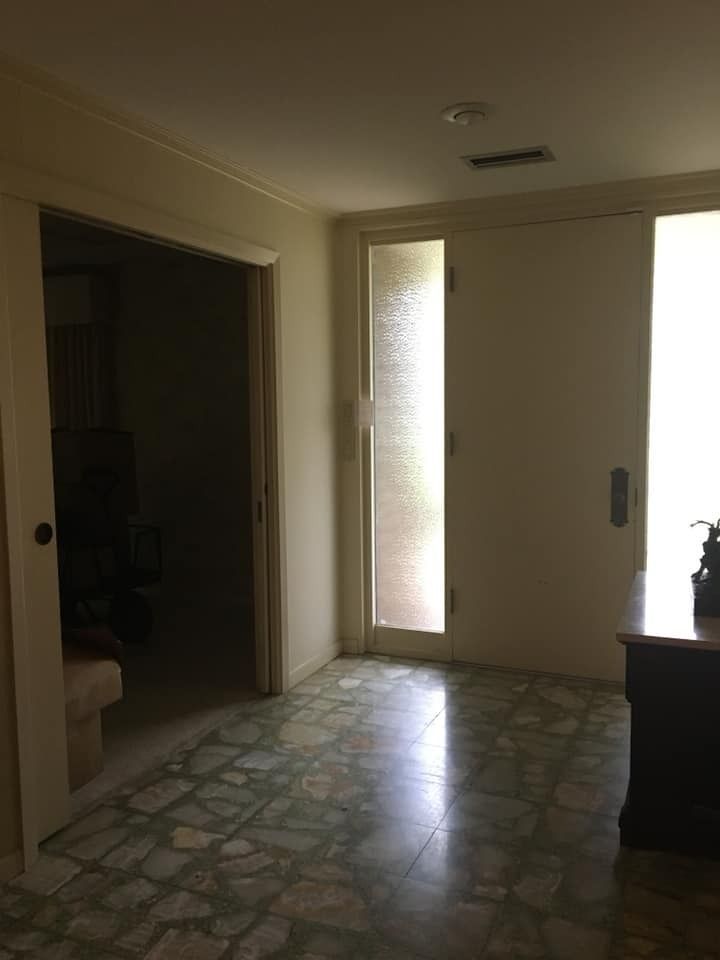 A sunlit foyer with a textured tile floor, featuring a front door with a tall side window and an adjacent open doorway.