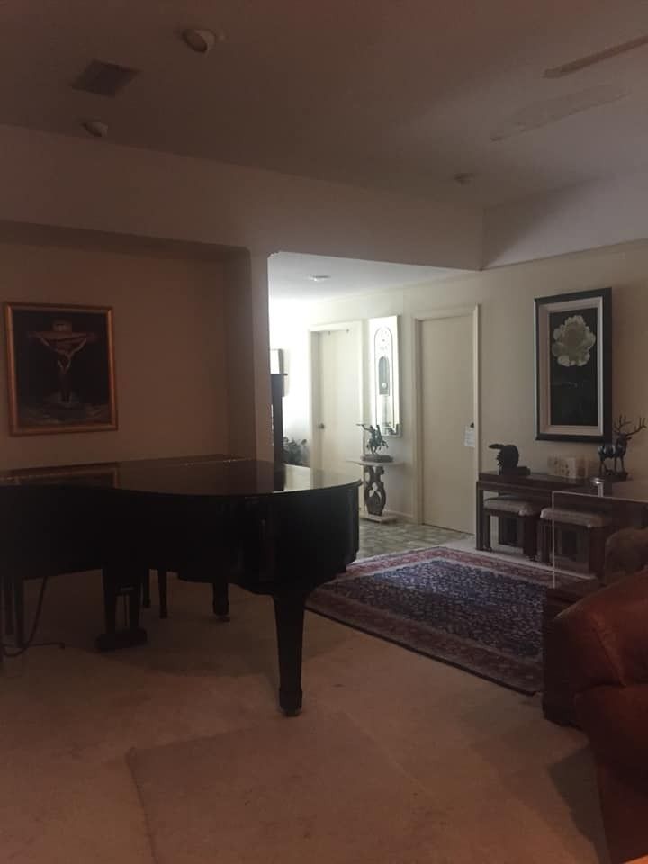 A grand piano sits in the foreground of a dimly lit living room with framed artwork, a patterned rug, and hallway doors.