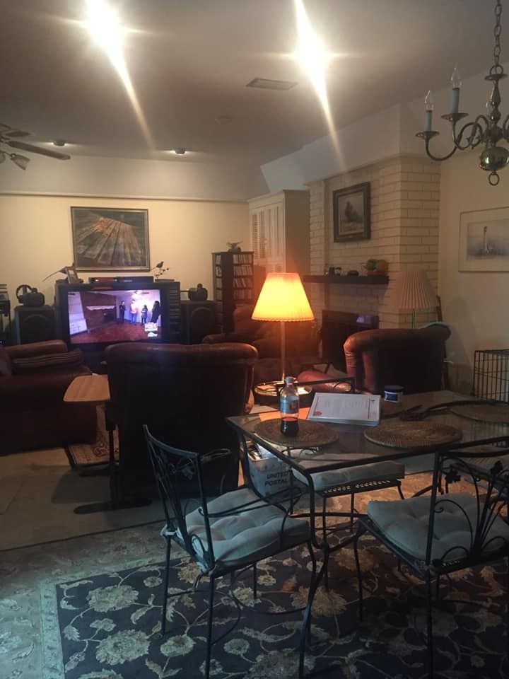 A cozy living and dining area with a glowing lamp on a glass table, dark chairs, a TV, and a fireplace in the background.