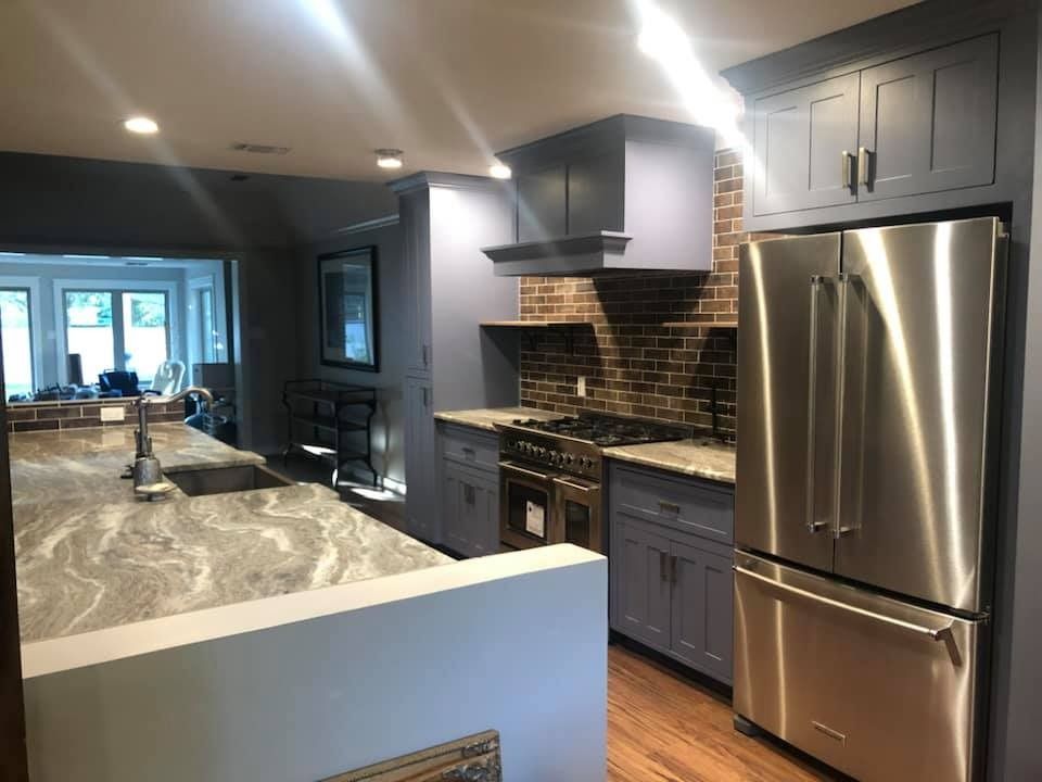 A modern kitchen with grey cabinetry, a stainless steel refrigerator, patterned countertops, and a brick backsplash.