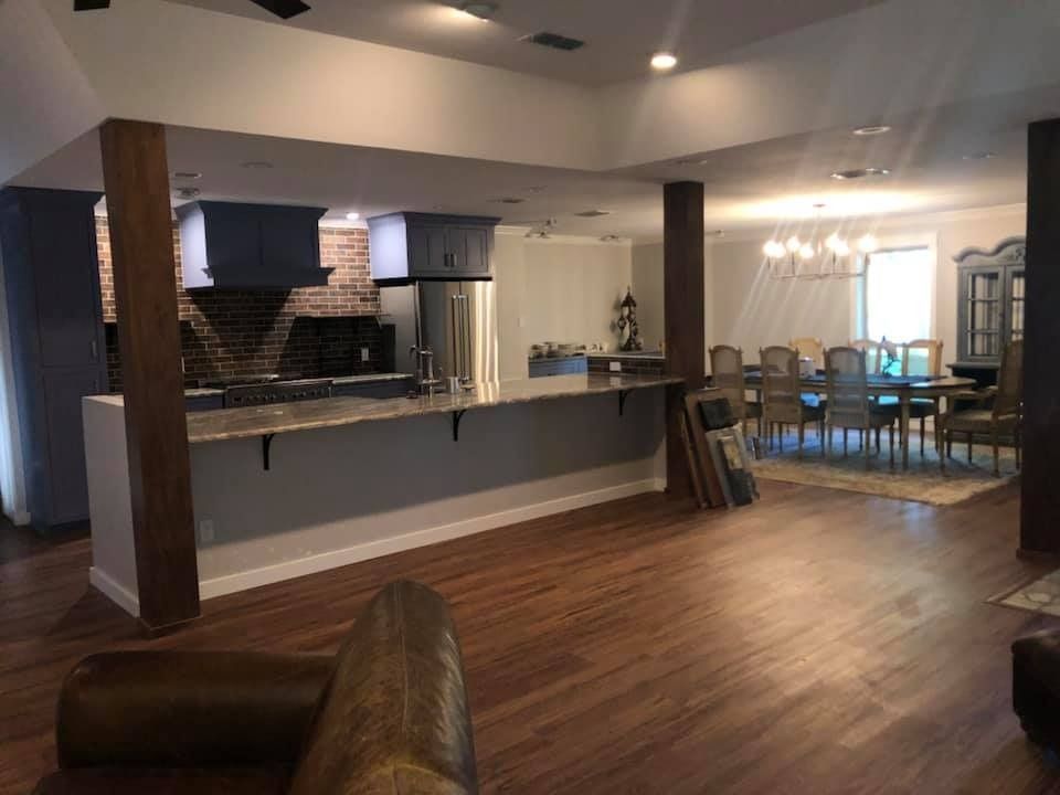 An open-concept room features a kitchen with a gray island and brick backsplash, leading to a dining area with a chandelier.