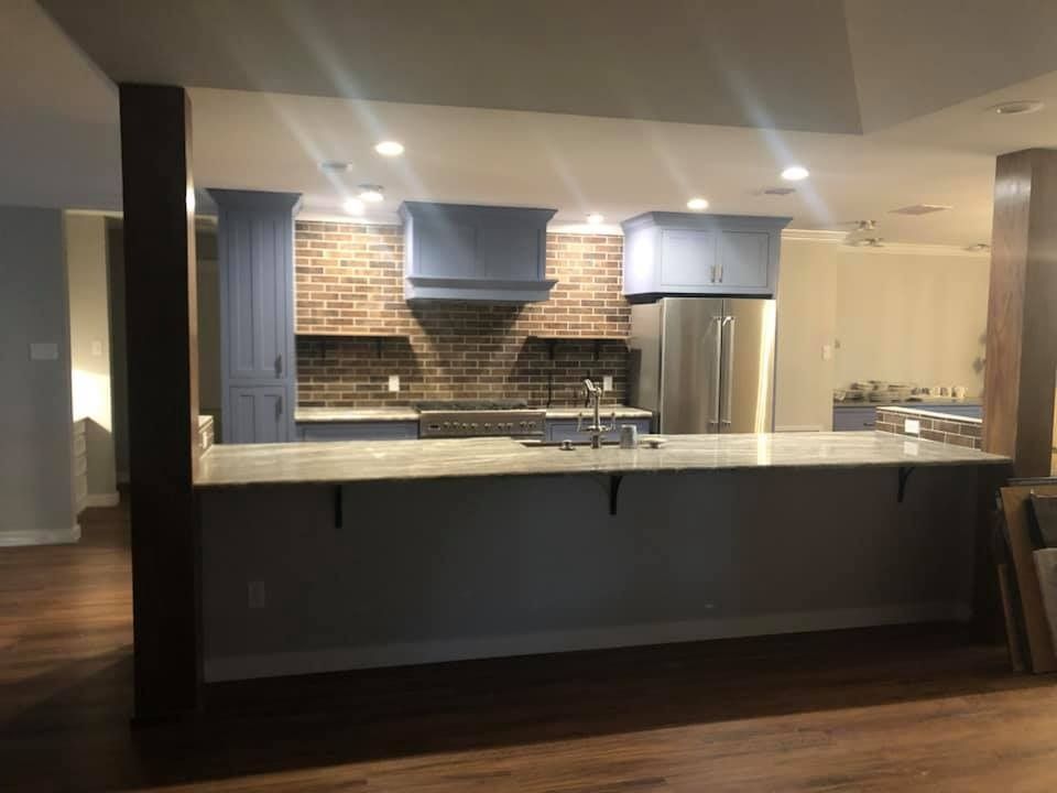 A kitchen island with a light countertop in front of blue cabinets, a brick backsplash, and stainless steel appliances.
