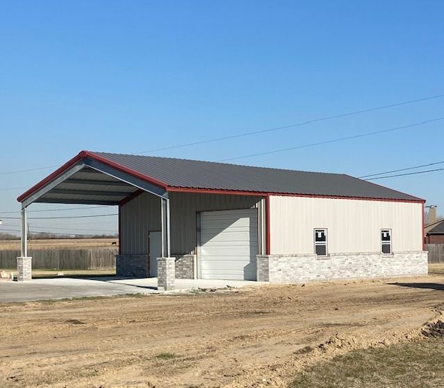 A tan metal building with a dark roof and stone base features an attached open-air carport under a clear blue sky.