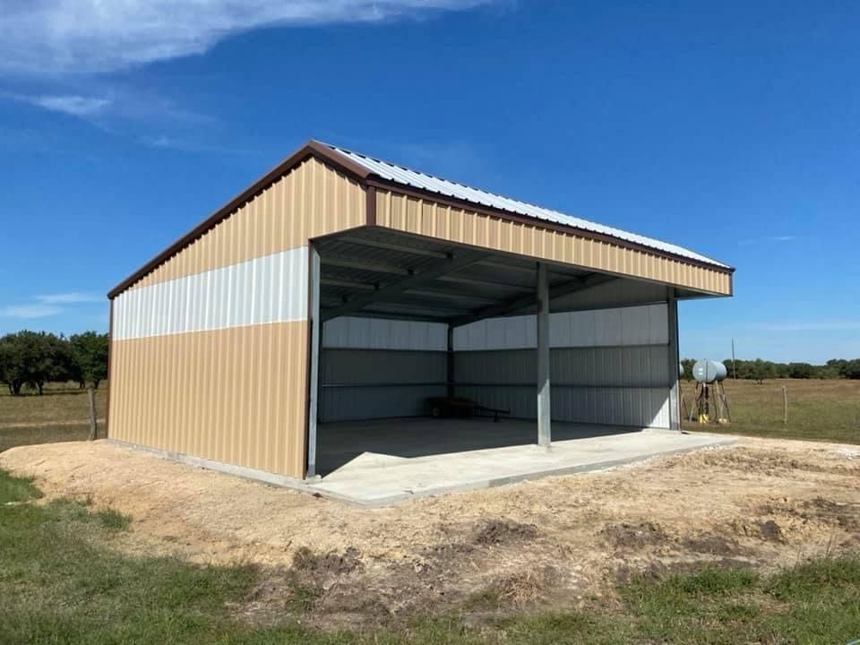 A metal pole barn with tan and white siding on a concrete slab in a grassy field under a bright blue sky.