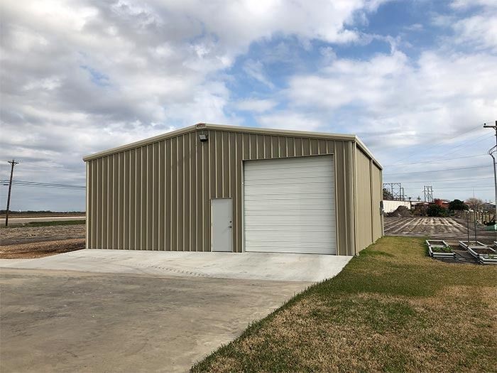 Tan metal warehouse building with a large rolling door and a single pedestrian door, set on a concrete pad under a sky.