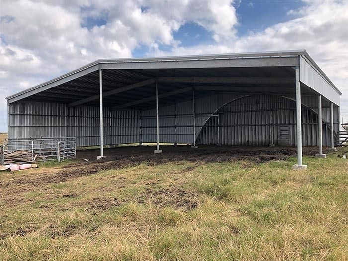 A large, open-sided steel farm shed with a corrugated metal roof, standing on dirt and grass under a cloudy sky.
