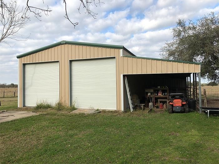 Tan metal workshop with two closed roll-up doors and an open-sided carport on the right, set in a grassy rural landscape.