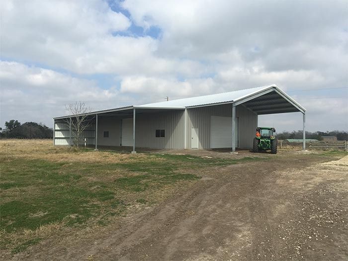 A light-colored metal barn with a covered carport area, situated in a grassy field with a tractor parked underneath.