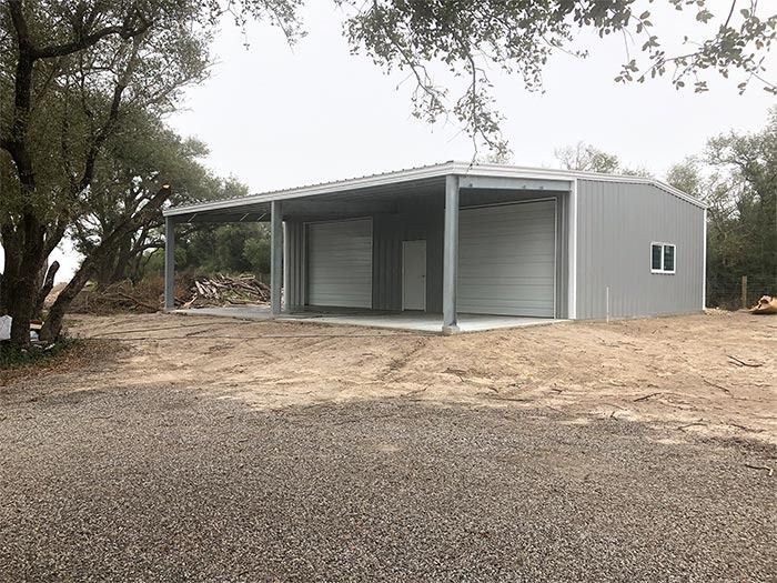 A grey metal building with an open-sided carport, two garage doors, and a pedestrian door, set on a gravel lot near trees.