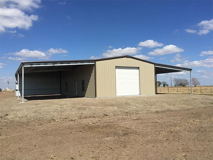 A tan metal barn with a roll-up garage door and open-sided overhangs stands on a flat, dry dirt lot under a blue sky.