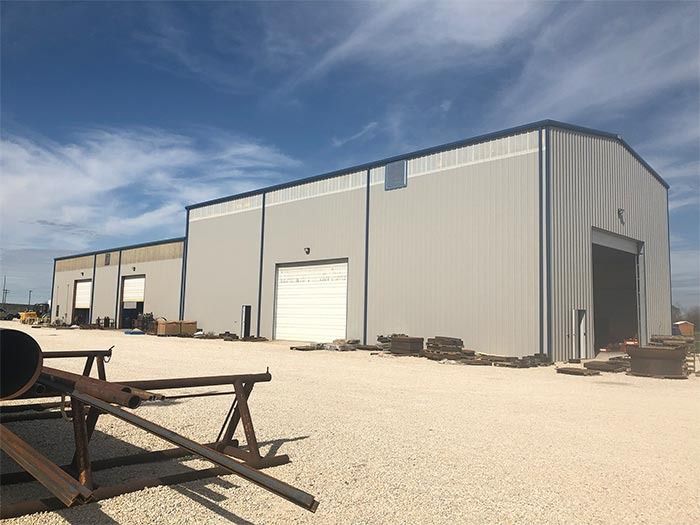 A large, beige metal industrial warehouse with multiple garage doors on a gravel lot under a blue sky.