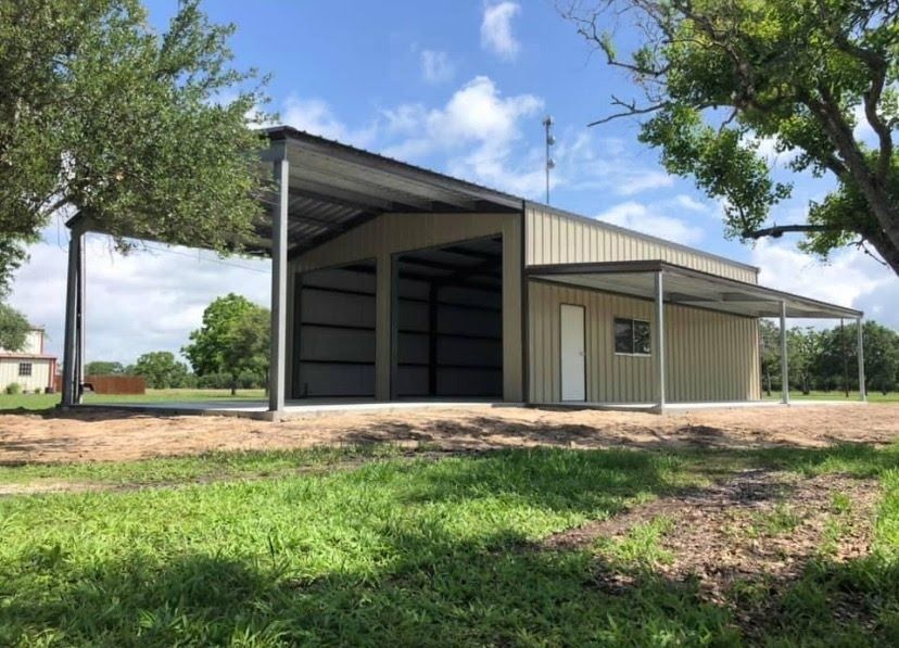 A beige metal building with an attached carport, positioned on a grassy lot with trees under a blue sky.