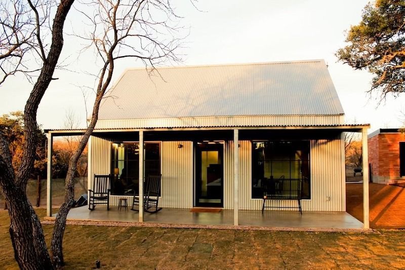 A beige metal cabin with a white gabled roof and a covered front porch featuring two rocking chairs under a sunny sky.