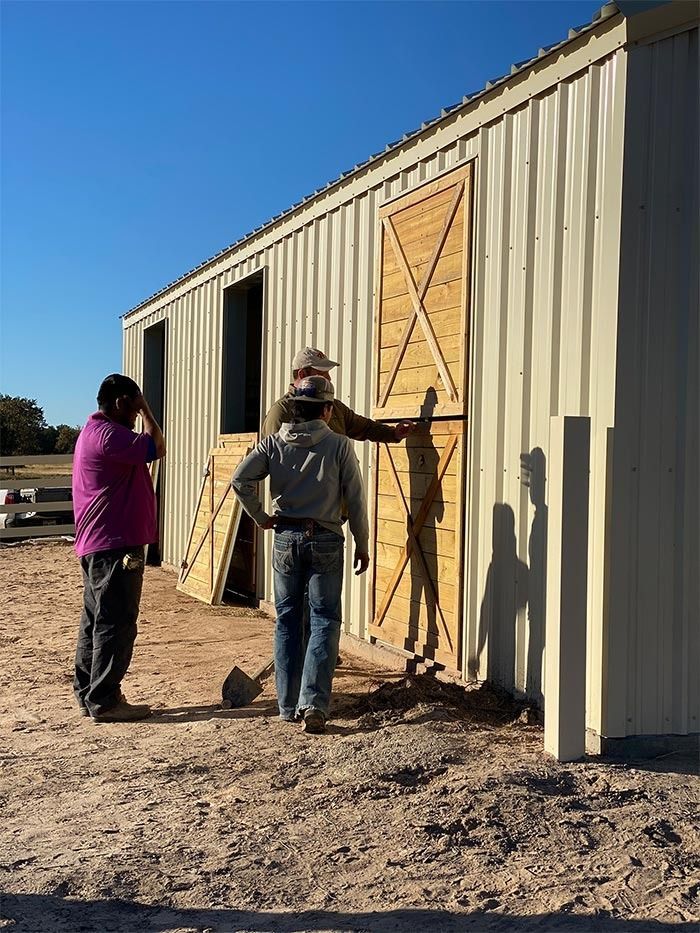 Two people stand outside a tan metal barn, looking at wooden stall doors during construction on a sunny, clear day.
