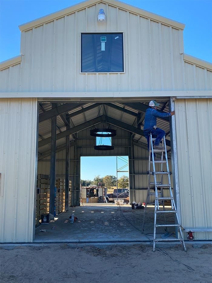 A person on a ladder installs a metal trim piece on the frame of a large, beige, open-front metal barn.