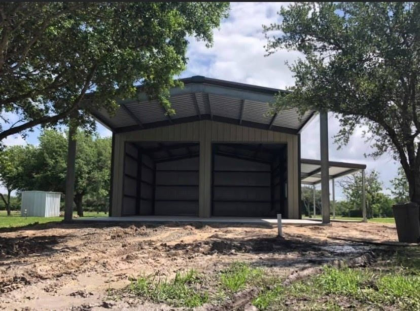 A tan, open-front metal garage with a dark roof and side porch, situated on a dirt lot surrounded by trees.