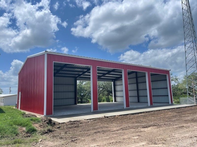 A red metal-sided, three-bay open garage structure under a blue sky with white clouds.