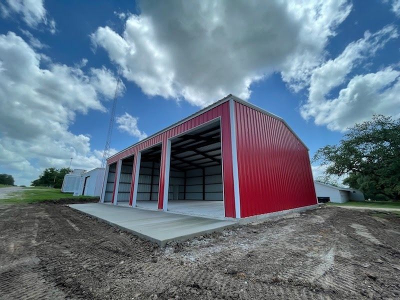 A red metal-sided barn with two large open bays sits on a fresh concrete pad under a blue sky with white clouds.