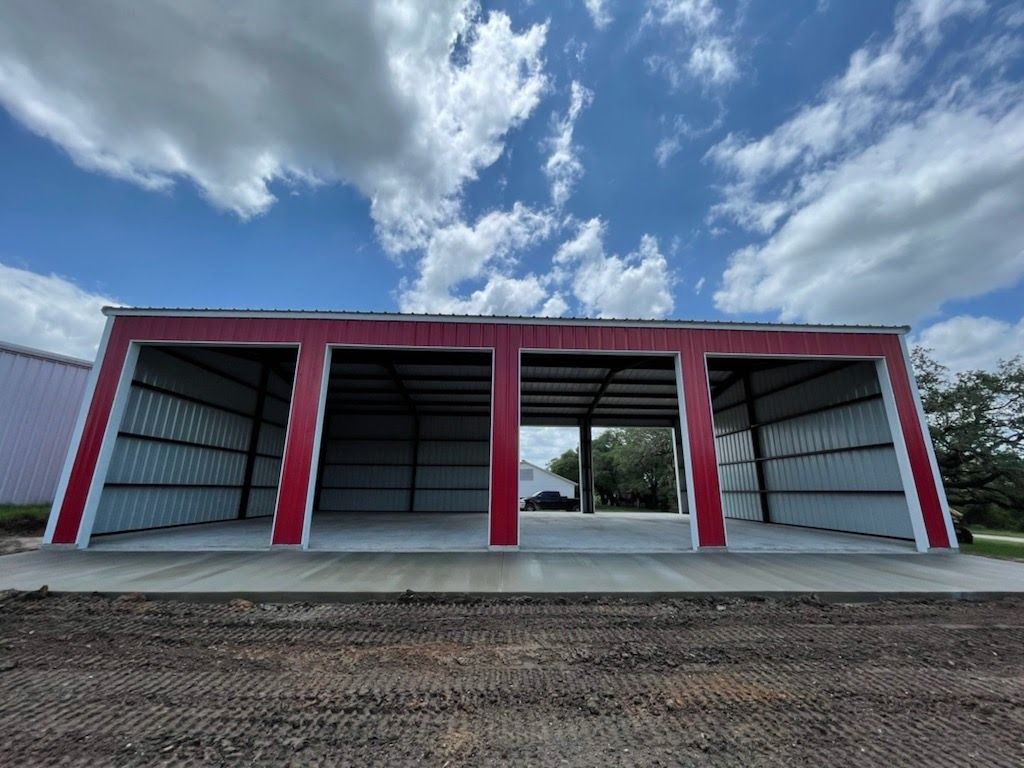A wide, open-sided metal carport with a red frame and white siding, set on a concrete slab under a blue sky.