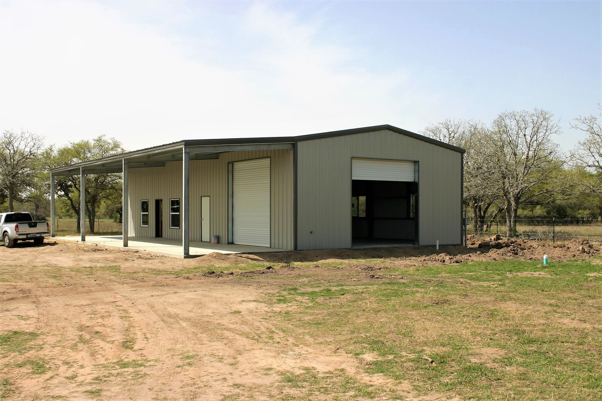 A light gray metal workshop building with a side porch, a roll-up door, and a white pickup truck parked outside.