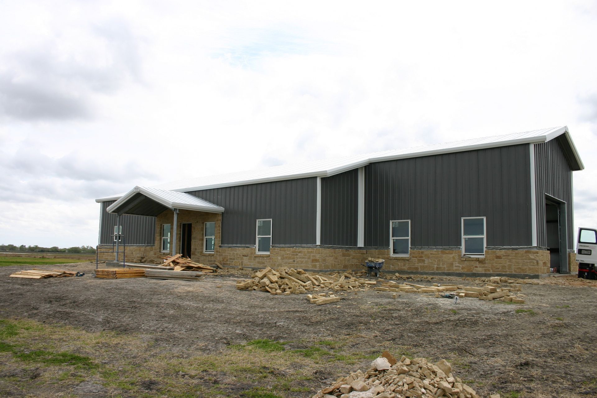 A newly constructed metal building with gray siding, a tan stone base, and a front entrance canopy on a dirt lot.