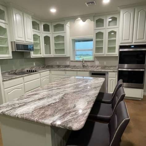 A modern kitchen featuring white cabinets, a large granite island with dark gray barstools, and stainless steel appliances.