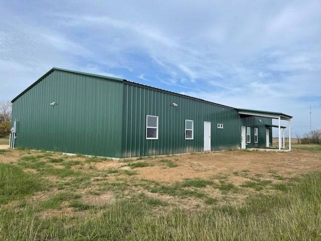 A large, rectangular green metal building with a white trim and doors sits on a dirt lot under a blue sky.