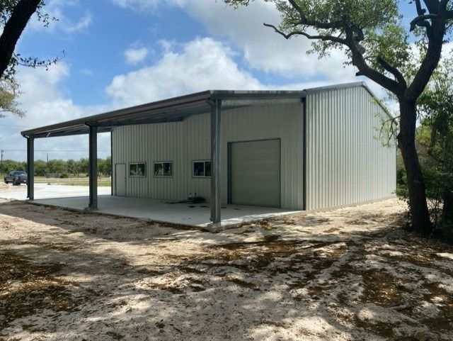 A light-colored metal workshop building with an open-sided carport, situated on a dirt lot surrounded by trees.