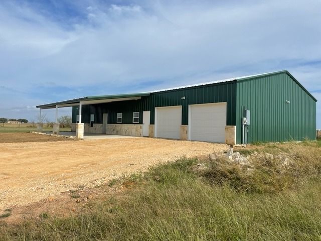 A green metal building with two white garage doors, a stone base, and a side porch on a gravel lot under a blue sky.