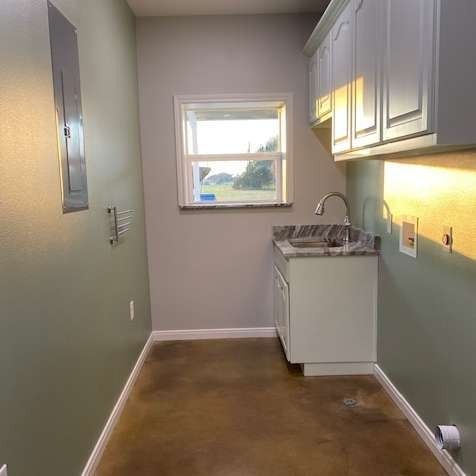 A small utility room with green walls, a white sink cabinet, overhead cupboards, a window, and a concrete floor.