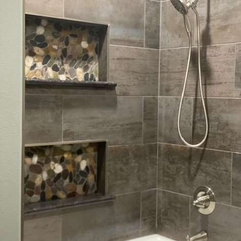 A shower with large, dark rectangular wall tiles and two recessed wall niches filled with multicolored river stones.