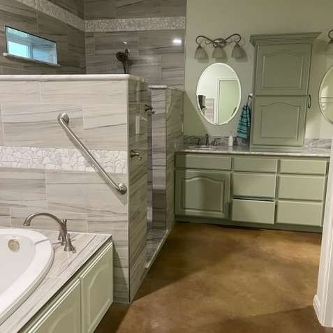 A bathroom with a jetted tub, a tiled walk-in shower with a grab bar, and sage green cabinetry under a circular mirror.