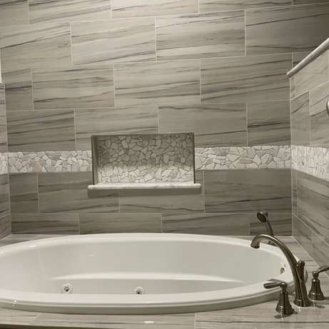 A white oval soaking tub with a silver faucet set against gray stone-tiled walls with a decorative mosaic inset shelf.