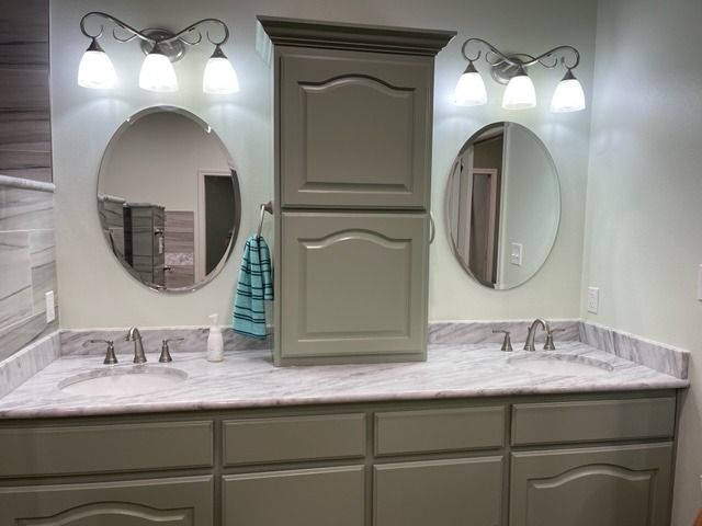 A sage green bathroom vanity with double sinks, marble countertop, twin oval mirrors, and a central storage cabinet.
