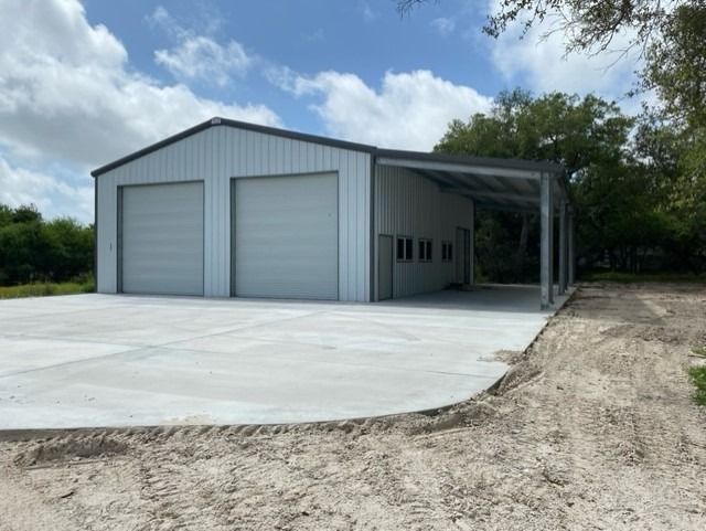 A light gray metal warehouse with two garage doors and an open-sided carport, set on a large concrete pad.