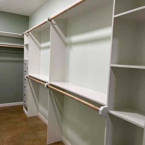 A walk-in closet with white wooden shelving and natural wood hanging rods against light green walls and wood flooring.