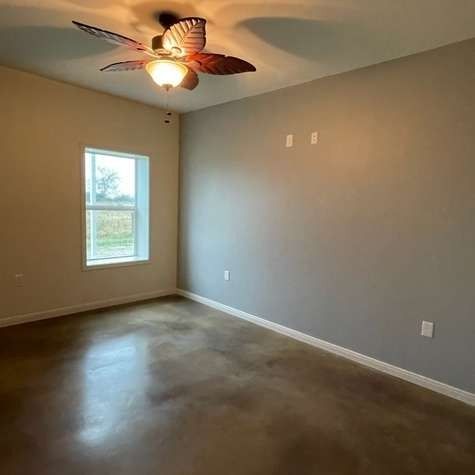 A bright, empty room with grey walls, polished concrete floors, a window on the left, and a ceiling fan in the center.