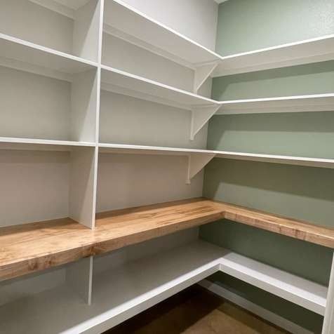 A walk-in pantry with white wooden shelves, a natural wood countertop, and sage green walls.