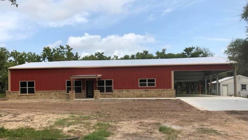 A modern red metal barn-style building with stone skirting, a front porch, and an open side garage under a clear sky.