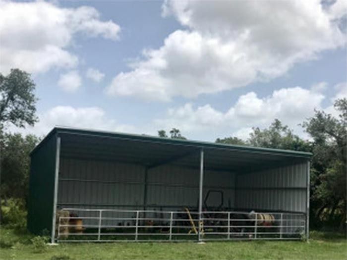 A dark green open-sided metal shed with a white interior sits in a grassy field under a cloudy blue sky.