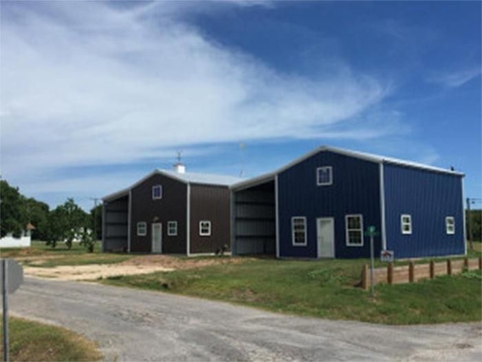 A brown building and a blue building stand side by side on a grassy lot next to a dirt road under a blue sky.