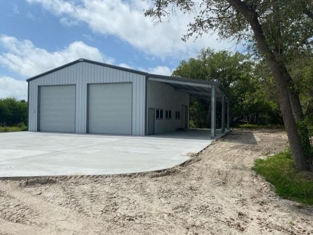 A light gray metal garage with two roll-up doors and an attached side carport, set on a concrete pad with dirt surroundings.