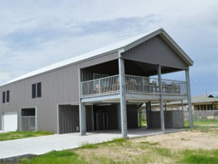 A gray, two-story metal building with a raised deck, concrete driveway, and open lower level on a clear day.