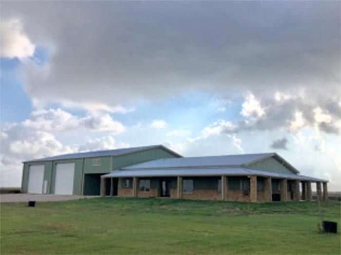 A green metal building with a wrap-around porch and two garage bays in a grassy field under a cloudy, blue sky.