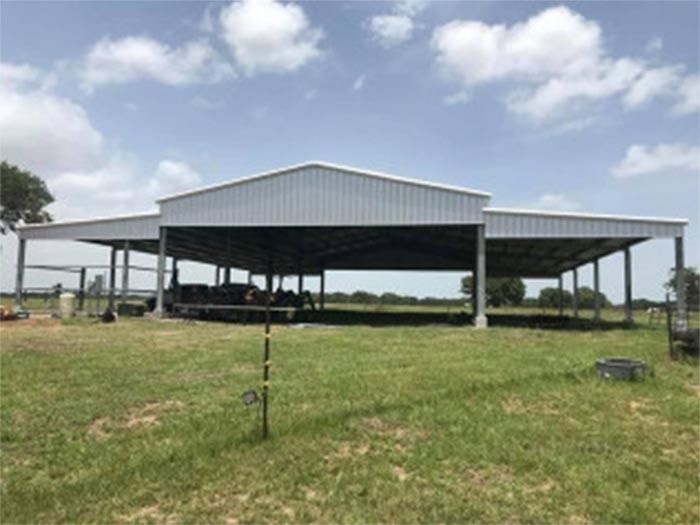 A wide metal pole barn with a gabled roof in a grassy field under a bright, cloudy sky.